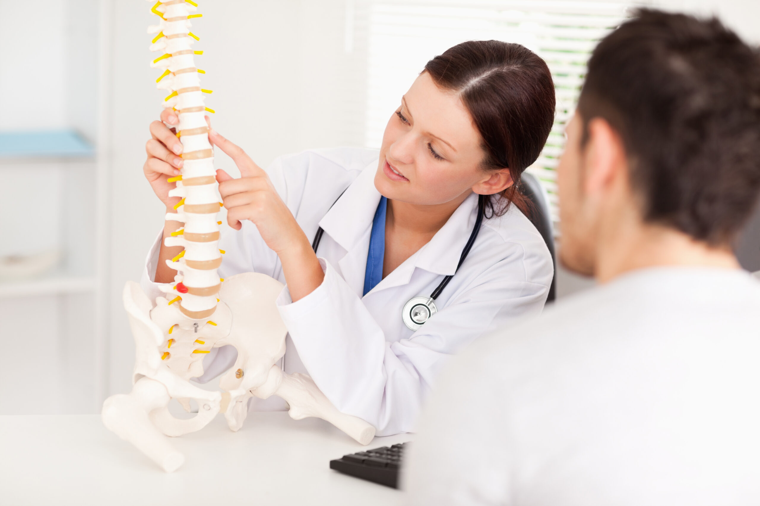 A female back pain doctor holding a model of the human spine and speaking with a male patient about his treatment options.
